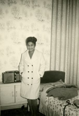A young woman wearing a smart skirt suit stands next to a bed